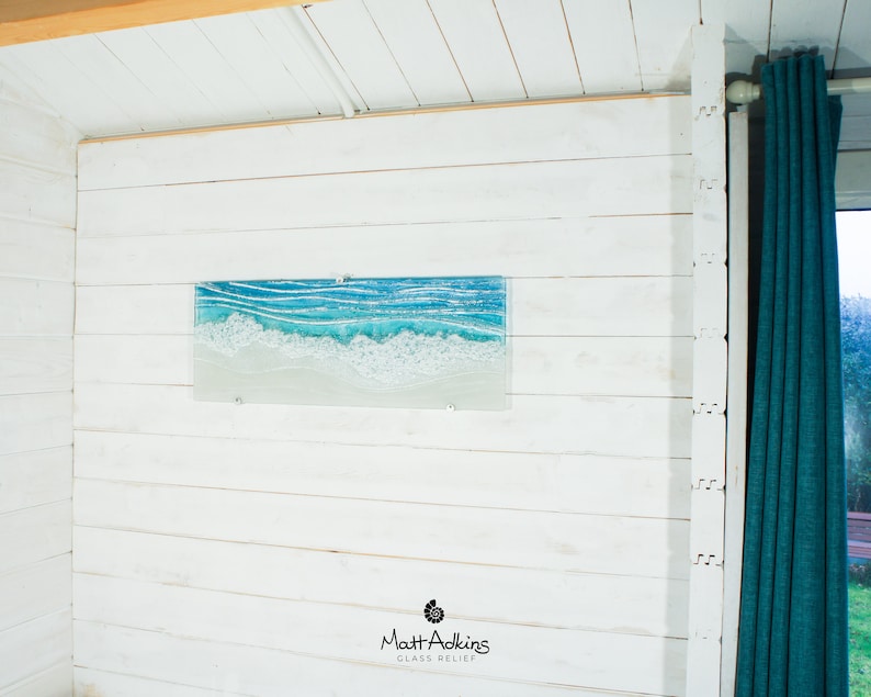 Puede incluir: Una pared de madera blanca con una pieza de arte de vidrio azul y blanco que representa olas. La obra de arte se titula "Matt Adkins Glass Relief".