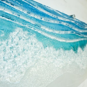 Puede incluir: Primer plano de una obra de arte de vidrio azul y blanco que representa una ola rompiendo en la costa. La ola est&aacute; hecha de vidrio en capas con una cresta de espuma blanca texturizada.