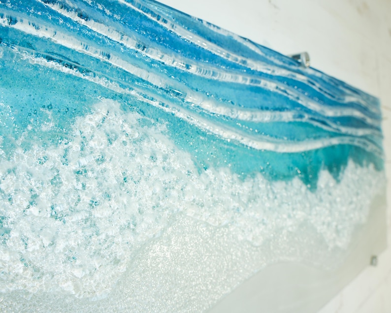 Puede incluir: Primer plano de una obra de arte de vidrio azul y blanco que representa una ola rompiendo en la costa. La ola est&aacute; hecha de vidrio en capas con una cresta de espuma blanca texturizada.