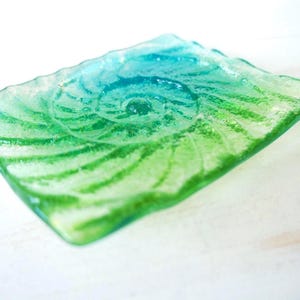 Puede incluir: Un plato rectangular de vidrio con un degradado vibrante de tonos verdes y azules. La superficie presenta un patrón en espiral texturizado y en relieve que recuerda a una concha de amonita, con bordes ondulados y un acabado translúcido.