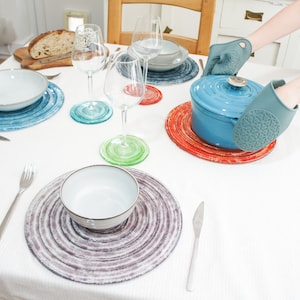 Puede incluir: Una olla de cerámica azul con tapa se coloca sobre un posavasos de color rojo y naranja con un patrón de espiral. La olla está siendo sostenida por una mano que lleva un guante de horno de silicona gris. La olla está sobre un mantel blanco con otros posavasos y platos.