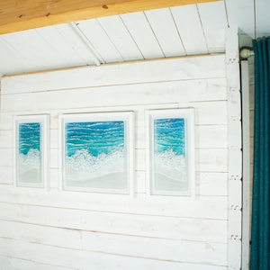 May include: Three framed pieces of art depicting a blue ocean scene with white foamy waves. The frames are white and the wall is white wood panelling.