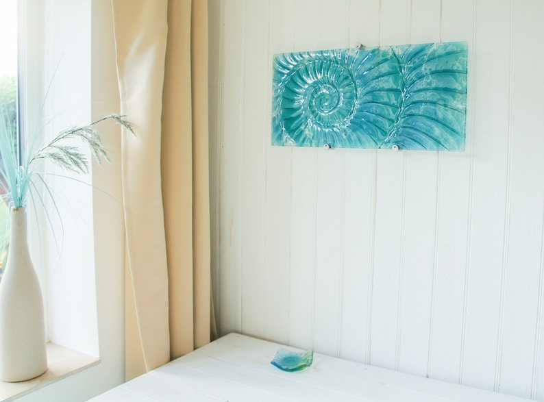 May include: A teal blue glass wall art piece featuring a spiral design. The art is mounted on a white wall with white vertical paneling.