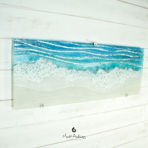 Puede incluir: Una pieza rectangular de arte de pared de vidrio que representa un paisaje mar&iacute;timo con olas azules y blancas rompiendo en una playa de arena. La pieza est&aacute; montada en una pared de madera blanca.