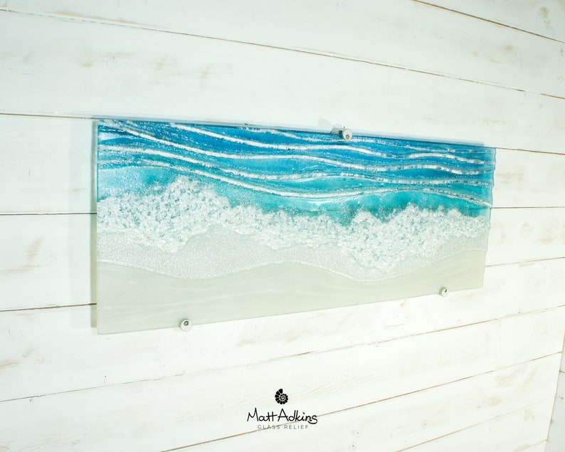 Puede incluir: Una pieza rectangular de arte de pared de vidrio que representa un paisaje mar&iacute;timo con olas azules y blancas rompiendo en una playa de arena. La pieza est&aacute; montada en una pared de madera blanca.