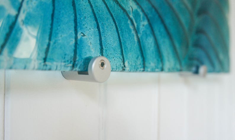 May include: Close-up of a silver wall mount with a screw visible. The mount is attached to a piece of blue glass art.