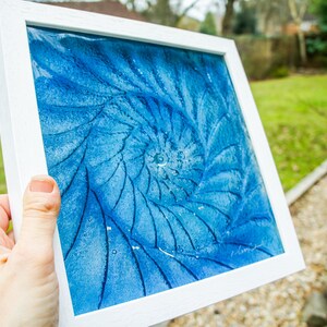 May include: A square artwork in a white frame featuring a deep blue spiral pattern resembling a nautilus shell. The surface has a textured, granular appearance with darker blue lines radiating from the center, creating a dynamic, swirling effect.