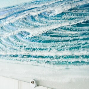 May include: A close-up of a glass art piece depicting ocean waves. The waves are made of layered glass with white accents to represent foam. The piece is mounted on a white tiled wall.