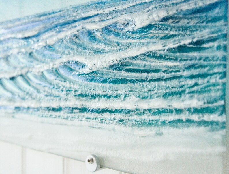 May include: A close-up of a glass art piece depicting ocean waves. The waves are made of layered glass with white accents to represent foam. The piece is mounted on a white tiled wall.