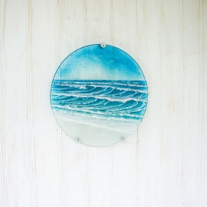 May include: A circular fused glass wall art piece depicting a coastal ocean scene. The design features a gradient of blue sky, textured white-capped waves rolling toward the shore, and a smooth, light-colored sandy beach at the bottom edge.
