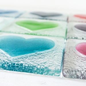 Puede incluir: Una colección de posavasos cuadrados de vidrio, cada uno con una forma de corazón en relieve en un color diferente, incluyendo azul, verde, rosa y morado. El vidrio tiene un acabado texturizado y burbujeante, y los corazones son lisos.