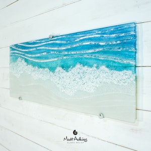 Puede incluir: Una pieza de arte de pared rectangular de vidrio que representa una escena de oc&eacute;ano azul con olas blancas espumosas rompiendo en una playa de arena. La pieza est&aacute; montada en una pared blanca con soportes plateados.