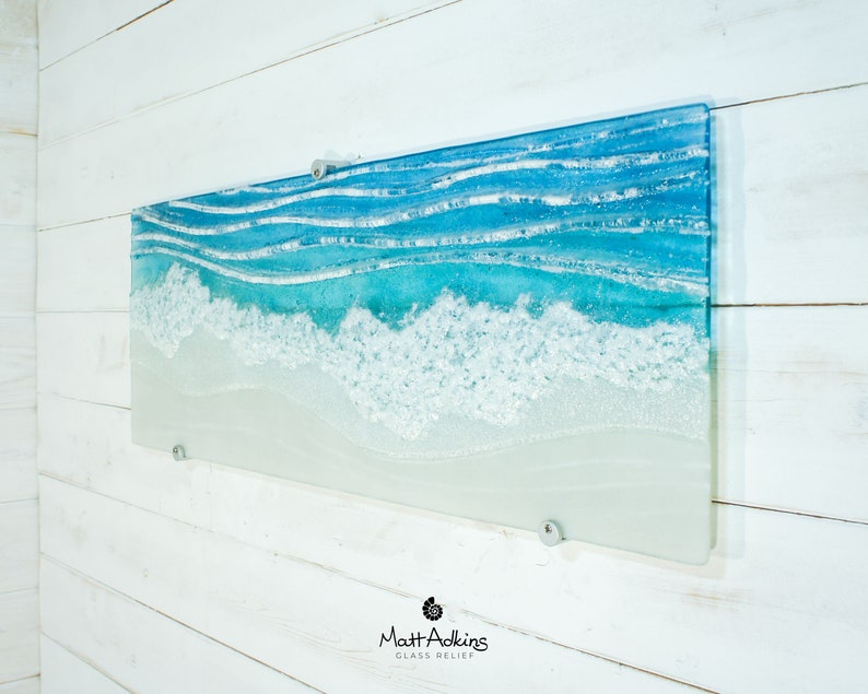 Puede incluir: Una pieza de arte de pared rectangular de vidrio que representa una escena de oc&eacute;ano azul con olas blancas espumosas rompiendo en una playa de arena. La pieza est&aacute; montada en una pared blanca con soportes plateados.