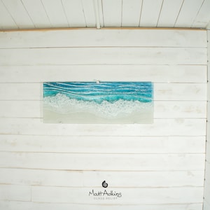 Puede incluir: Una pieza de arte de vidrio rectangular que representa una escena de oc&eacute;ano azul con olas blancas espumosas rompiendo en una playa de arena. La pieza est&aacute; montada en una pared de madera blanca. El nombre del artista, "Matt Adkins", y las palabras "Glass Relief" est&aacute;n impresas debajo de la obra de arte.