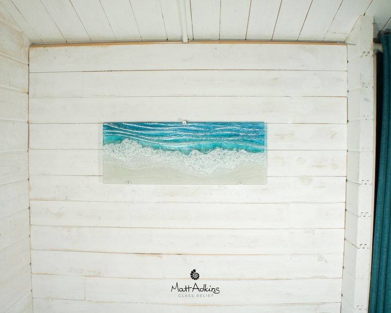 Puede incluir: Una pieza de arte de vidrio rectangular que representa una escena de oc&eacute;ano azul con olas blancas espumosas rompiendo en una playa de arena. La pieza est&aacute; montada en una pared de madera blanca. El nombre del artista, "Matt Adkins", y las palabras "Glass Relief" est&aacute;n impresas debajo de la obra de arte.