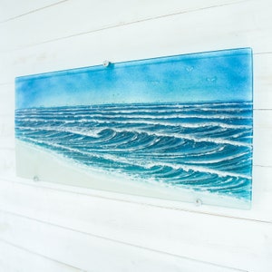 May include: A rectangular fused glass wall art piece depicting a coastal ocean scene. The artwork features a light blue sky, deep blue rolling waves with white foam crests, and a smooth, pale sandy beach along the bottom edge of the glass panel.