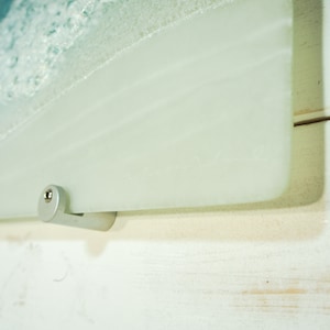 Puede incluir: Primer plano de un soporte de pared de metal plateado fijado a un panel de vidrio esmerilado. El soporte tiene un peque&ntilde;o tornillo en el lateral.