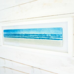 May include: A rectangular wall art piece featuring a fused glass panel depicting a coastal scene with a sandy beige shore, multiple horizontal bands of blue ocean waves, and a light blue sky, all encased within a simple white wooden frame.