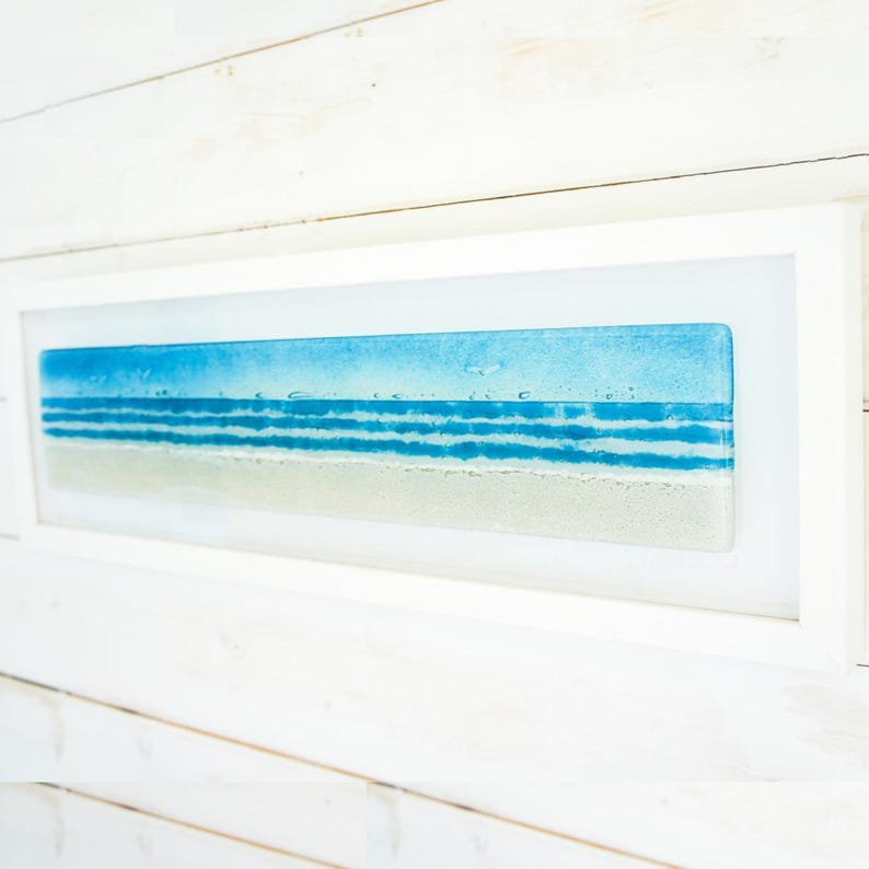 May include: A rectangular wall art piece featuring a fused glass panel depicting a coastal scene with a sandy beige shore, multiple horizontal bands of blue ocean waves, and a light blue sky, all encased within a simple white wooden frame.