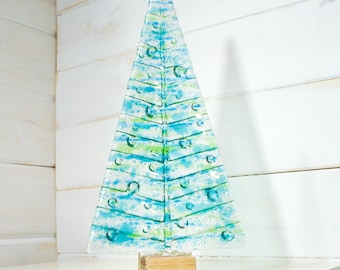 May include: A decorative glass Christmas tree with a triangular shape. The tree features a mix of blue and green colors with bubble-like accents. It is mounted on a small, rectangular wooden base, perfect for holiday decor.