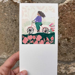 Peut inclure: Une impression d'art rectangulaire représentant une personne à vélo rose à travers un champ de fleurs roses. La personne porte une chemise violette et un pantalon vert. Le fond a un motif floral et le texte "Matild Gros".