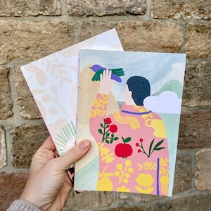 Può includere: Due biglietti di auguri tenuti in mano. Un biglietto presenta un'illustrazione colorata di una persona che tiene in mano un uccello, con una giacca a fantasia rosa e gialla. L'altro biglietto ha uno sfondo bianco con un motivo floreale.