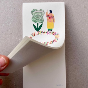 Peut inclure: Bloc-notes avec une illustration fantaisiste. L'œuvre représente une silhouette en manteau jaune avec des détails floraux, près d'une grande fleur verte et blanche. Un cercle coloré entoure la silhouette. Le nom de l'artiste, Matilda Dres, est visible.
