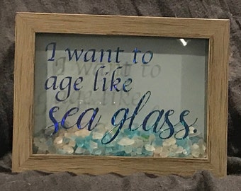 May include: A framed wooden sign with a blue and iridescent vinyl quote that reads "I want to age like sea glass." The frame contains a collection of sea glass.