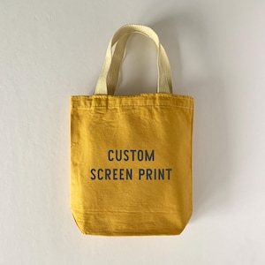 May include: A mustard yellow canvas tote bag with two light beige fabric handles. The front of the bag features the text CUSTOM SCREEN PRINT printed in a dark gray sans-serif font centered on the body of the bag.