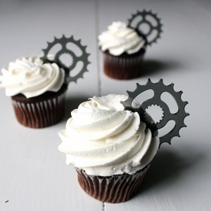 Könnte beinhalten: Drei Schokoladen-Cupcakes mit weißem Frosting und silbernen Zahnrad-Toppern.