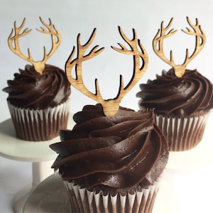 Puede incluir: Tres cupcakes de chocolate con envoltorios de papel blanco y adornos de madera con forma de cuernos de ciervo. Los cupcakes tienen un remolino generoso de glaseado de chocolate oscuro. Un cupcake está sobre un pequeño pedestal blanco.