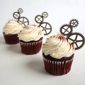 May include: Three red velvet cupcakes with white frosting and decorative metal gears on top.