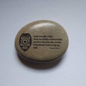 May include: A gray stone with a black and white police badge and a bible verse engraved on it. The verse reads "Keep me safe, LORD, from the hands of the wicked; protect me from the violent, who devise ways to trip my feet. Psalm 140:4"