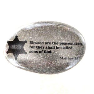 May include: A gray stone with a black sheriff's star and the quote "Blessed are the peacemakers, for they shall be called sons of God. Matthew 5:9"