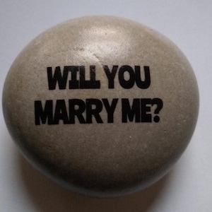 May include: A gray stone with the words "Will you marry me?" written in black lettering.