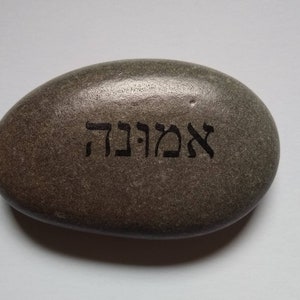 May include: A smooth, oval-shaped gray stone with the Hebrew word "Emuna" etched into its surface. The stone has a natural, textured appearance and is set against a plain white background. The engraved text is in a dark color.