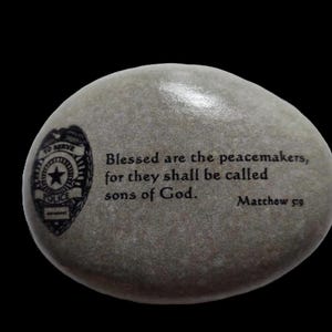 May include: A gray stone with a black and white police badge and a quote from the Bible: "Blessed are the peacemakers, for they shall be called sons of God. Matthew 5:9"