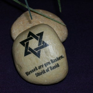 May include: Two smooth, beige stones. The front stone features a black Star of David symbol and the text "Blessed are you Hashem, Shield of David". The stones are on a dark purple surface.