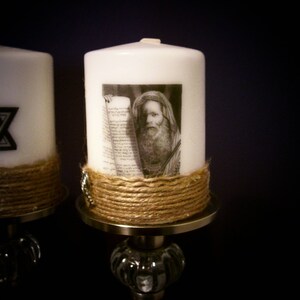 May include: White pillar candle featuring a black and white image of a man holding a scroll. The candle is wrapped with twine and sits on a decorative metal and glass candle holder. A second candle with a Star of David is visible.