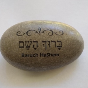 May include: A gray stone with a Hebrew inscription that reads "Baruch HaShem" which translates to "Blessed is the Name" in English. The stone is oval shaped and has a decorative flourish above the inscription.