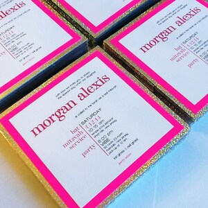 May include: A stack of invitations with a pink border and gold glitter. The invitations are for a bat mitzvah for Morgan Alexis. The text on the invitations includes the date, time, and location of the event.