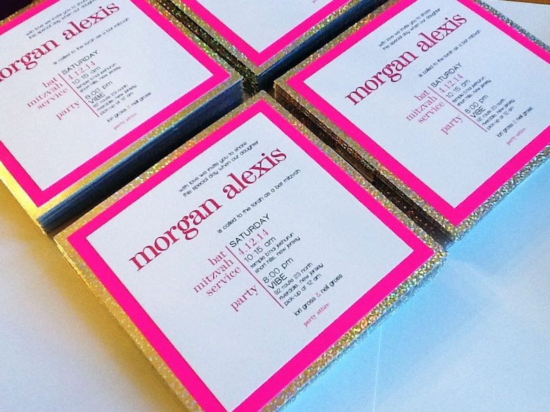 May include: A stack of invitations with a pink border and gold glitter. The invitations are for a bat mitzvah for Morgan Alexis. The text on the invitations includes the date, time, and location of the event.