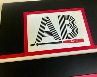 May include: A white card with a red border features the letters "AB" in black and white stripes. A black and red baseball bat is shown below the letters with the name "ALEX" printed on the bat.