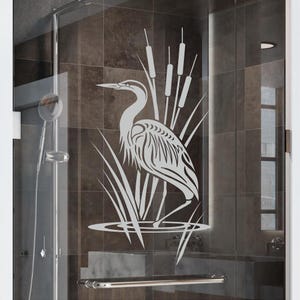 May include: A frosted glass shower door features a detailed heron design. The heron is depicted standing in water with reeds in the background. The design is in a light color, contrasting against the dark tile background.