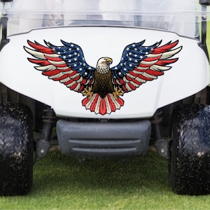 May include: A white golf cart with a large decal of an American bald eagle with outstretched wings. The eagle's wings are red, white, and blue, and the cart has black tires.