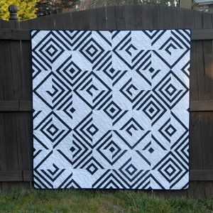 以下が含まれることがあります： 黒と白のキルトで、正方形と線の幾何学模様が施されています。キルトは木製のフェンスにかけられています。