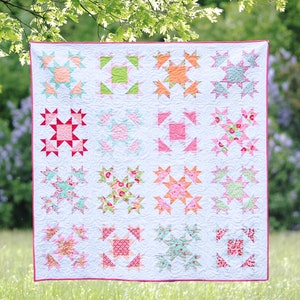 Puede incluir: Una colcha blanca con un patrón de estrellas en rosa, verde y naranja. La colcha está colgada de un tendedero en un campo de hierba.