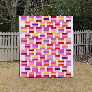 May include: A colorful quilt with a brick pattern in shades of pink, red, orange, yellow, and purple. The quilt is made with a white background and has a quilted texture.