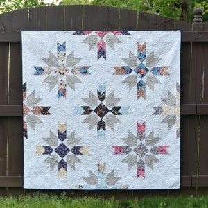 以下が含まれることがあります： グレー、ブルー、ピンクの星形の幾何学模様が施された白いキルト。キルトは茶色の木製フェンスにかけられています。