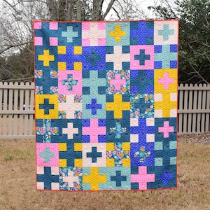 May include: A colorful quilt with a patchwork design featuring plus signs in various colors. The quilt has a red border and is displayed outdoors. Colors include teal, pink, yellow, blue, and white.